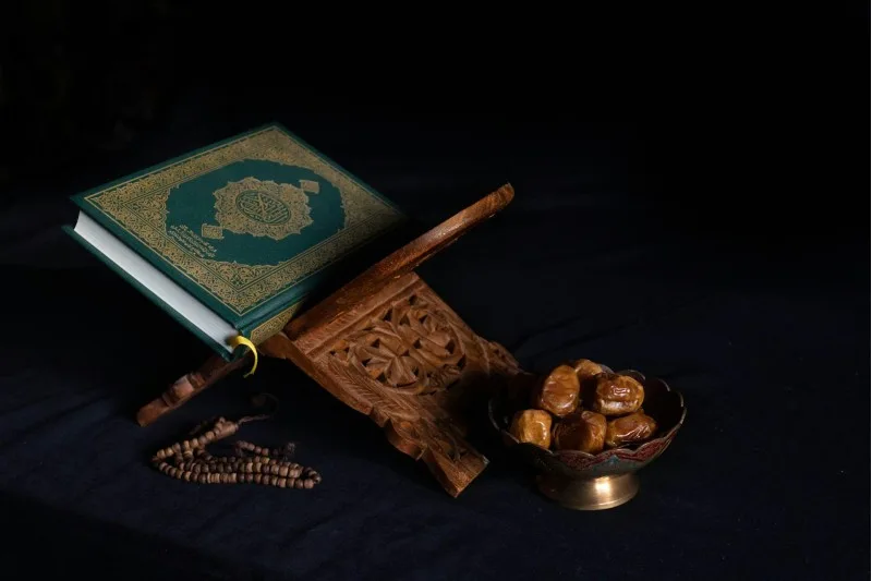 Oymalı ahşap rahle yanında yeşil kapaklı Kur'an-ı Kerim, tesbih ve hurma